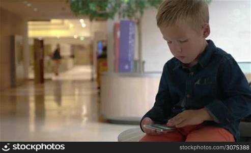 Little boy looking through the photos on smart phone in shopping mall, unidentified people passing by
