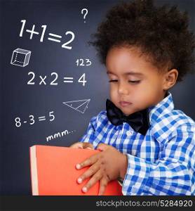 Little boy on math lesson, holding in hands book and standing near blackboard with written mathematical equation, back to school
