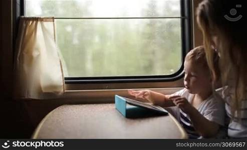 Little boy playing game on tablet PC sitting by window in the train. Mother helping him and they clapping hands when he wins