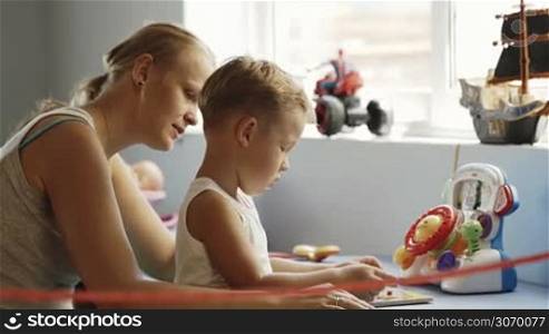 Little boy playing learning game at home, mother watching and kissing him