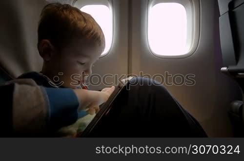 Little boy playing on tablet computer sitting by the illuminator in plane