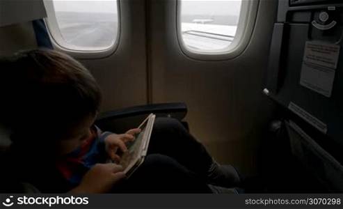 Little boy playing on touch pad in moving plane which going to take off. Traveling by air