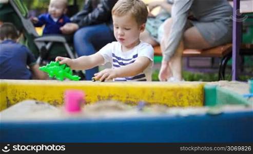 Little boy playing with plastic crocodile and car in the sand-pit in the yard. Outdoor activities of children. People with baby in background