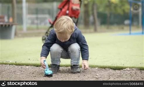 Little boy playing with toy digger outdoor and then going away. Playground with red baby carriage in background.