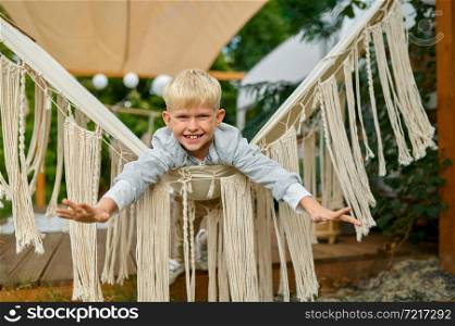 Little boy poses on hammock, summer camping. Family with children travel in camp car, nature and forest on background. Campsite adventure, travelling lifestyle, smiling kid camper. Little boy poses on hammock, summer camping