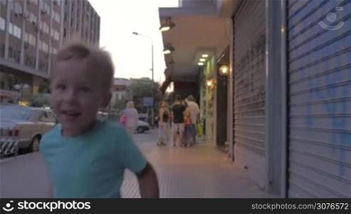 Little boy runs along a busy street of Thessaloniki, Greece. In the background seen a road, cars and shops