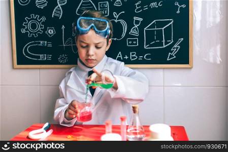 Little boy scientist pouring chemical green liquid from flask against of chalkboard with drawings. Serious kid playing with chemical liquids