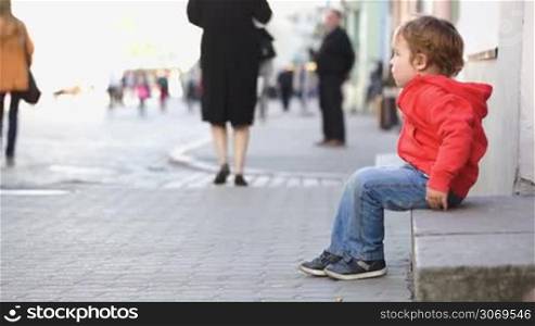 Little boy sitting alone in the crowded street. Defocus of people passing by in background