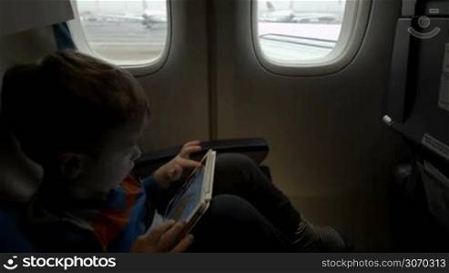 Little boy sitting in moving plane and playing on touch pad. Airplane is going to take off. Traveling by air