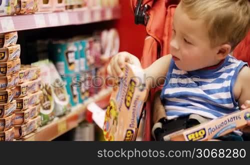Little boy sitting in the baby carriage and taking tasty things from shelf in the shop. He taking more and more.