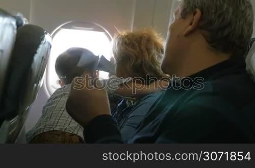 Little boy sitting on grandmothers lap and looking out plane window, grandfather trying to entertain him with paper plane. Family travel by air