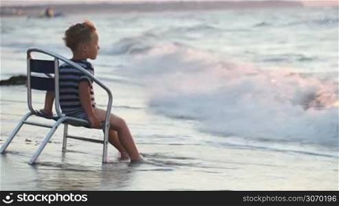 Little boy sitting on the beach on a windy day and watching how sea waves washing his feet
