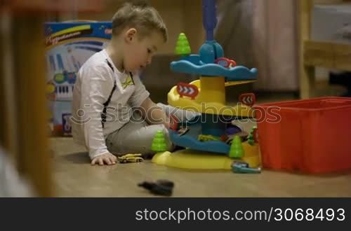 Little boy sitting on the floor in his playroom playing with a plastic parking garage and small miniature cars