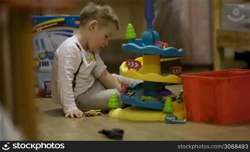Little boy sitting on the floor in his playroom playing with a plastic parking garage and small miniature cars