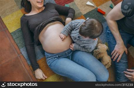 Little boy tickling the belly of his pregnant mother with his father watching. Toddler tickling the belly of his pregnant mother