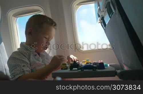 Little boy traveling by air. He sitting by the illuminator and playing with toy plane, many cars on the table in front of him. Entertaining during the flight