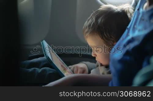 Little boy traveling in an airplane sitting in his seat playing with a tablet computer watched by a parent