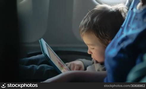 Little boy traveling in an airplane sitting in his seat playing with a tablet computer watched by a parent