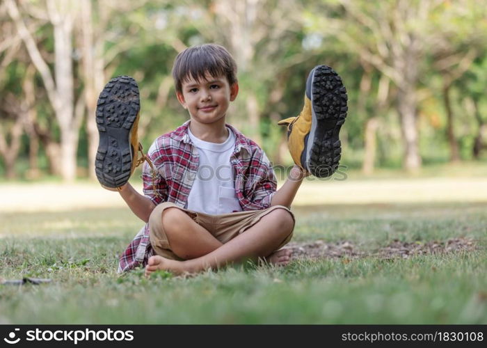 Little boy trying his father's hiking shoes sitting on green grass at park.