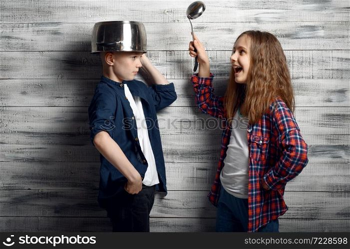 Little boy with a pot on his head and girl with the ladle in hand in studio. Children play, kids isolated on wooden background, child photo session. Kids with pot on head and ladle in hand