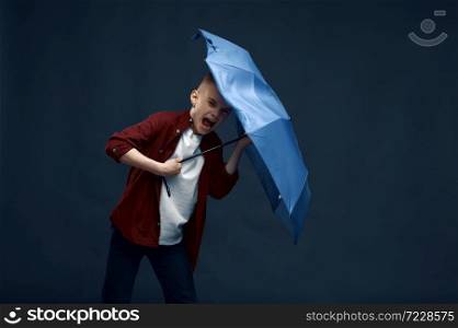 Little boy with a scared face holds an umbrella in studio, wind effect, powerful airflow. Children with developing hairs, kids isolated on dark background, child emotion. Boy with a scared face holds umbrella in studio
