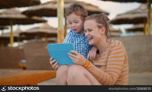 Little boy with his mother at a beach resort playing with a tablet computer on the beach under straw beach umbrellas