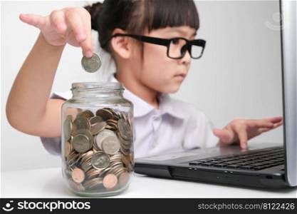 Little businesswoman puts a coin in a glass jar on a table while working with laptop in office. Children and business concept