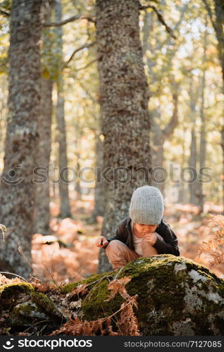 Little caucasian baby girl squatting wearing a wool cap in an autumn forest among ferns plays with plants