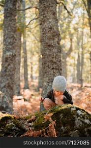 Little caucasian baby girl squatting wearing a wool cap in an autumn forest among ferns plays with plants