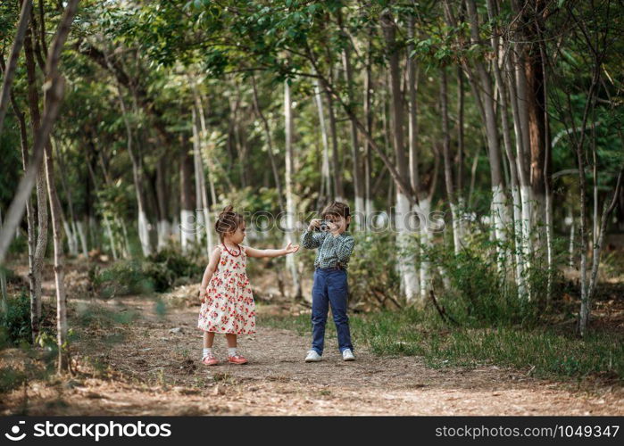 little Caucasian boy and girl in vintage clothes walking in the woods and the boy takes pictures of the girl on a retro camera. friendship and love of young children