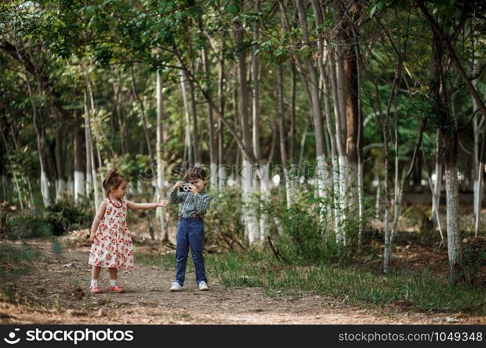 little Caucasian boy and girl in vintage clothes walking in the woods and the boy takes pictures of the girl on a retro camera. friendship and love of young children