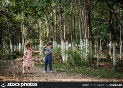little Caucasian boy and girl in vintage clothes walking in the woods and the boy takes pictures of the girl on a retro camera. friendship and love of young children