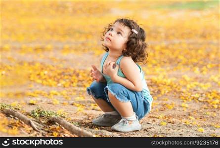 Little Caucasian child girl sitting under the tree, looking up to yellow leaves falling down on ground at park. Cute kid with curly hair wear casual clothes in Autumn. Copy space