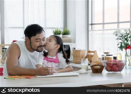 Little Caucasian cute daughter kissing cheek of Asian young father with beard while drawing with colored pencil together at home with glass of milk, marshmallows, breads and apples on kitchen table