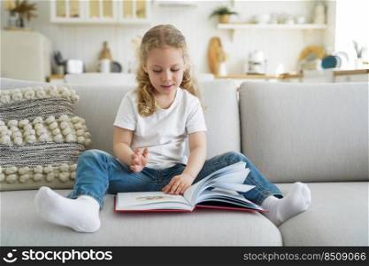 Little caucasian girl is sitting on couch and reading fairy tale book. Kid is studying at home on quarantine. Preschool child is learning to read. Leisure and study at cozy home on quarantine.. Little girl is sitting on couch and reading fairy tale book. Preschool child is learning to read.