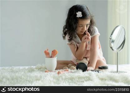 Little Caucasian kid girl holding eyebrow brush to apply color to eyebrows and eyelash looking at mirror with joy on white fluffy carpet. Cute child playing make up cosmetics with bear doll