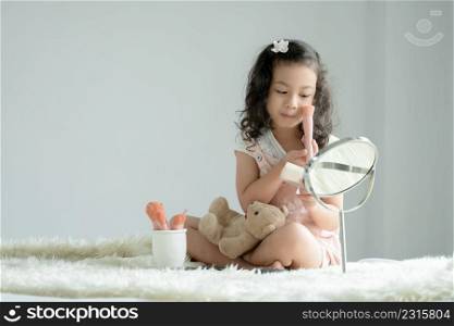 Little Caucasian kid girl holding pink blush brush to apply loose powder to cheek and face looking at mirror with joy on white fluffy carpet. Cute child playing make up cosmetics with bear doll