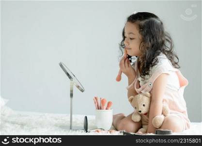Little Caucasian kid girl holding pink blush brush to apply powder to cheeks and face looking at mirror with joy on bed at home. Cute child playing make up cosmetics with bear doll. Side view