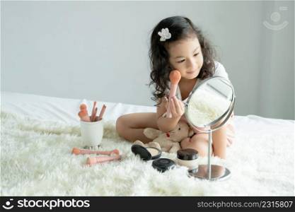 Little Caucasian kid girl holding pink blush brush to apply powder to cheeks and face looking at mirror with joy on bed at home. Cute child playing makeup cosmetics with bear doll. Copy space