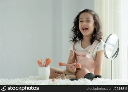 Little Caucasian kid girl playing makeup cosmetics sitting with bear doll. Cute child smiling and holding pink blush brush with joy and happiness on bed at home. Copy space