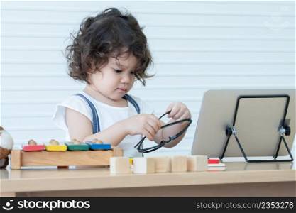 Little Caucasian nerd kid girl holding glasses while online learning on tablet at home with wooden block toys and colorful xylophone. White background