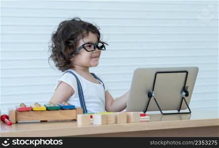 Little Caucasian nerd kid girl wearing glasses smiling while online learning on tablet at home and playing wooden block toys with colorful xylophone and puzzle pieces. White background