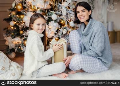Little child and her mother sit on warm white carpet near decorated New Year tree, holds present in hands, have pleased and happy expressions, glad to spend holidays together. Childhood and holidays