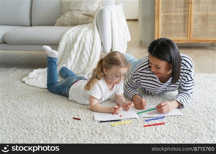 Little child daughter and young mom drawing painting by colorful pencils lying on floor carpet together. Mother teaches kid girl to draw. Educational pastime, children’s education concept.. Kid girl daughter and mom drawing painting lying on floor carpet together. Children’s education