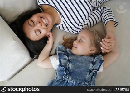 Little child daughter tickling foster mother, lying on couch at home. Top view of laughing young mom and adopted small preschooler girl having fun together, playing on cozy sofa. Happy motherhood.. Little child daughter tickling laughing mother, lying on sofa, having fun together. Happy motherhood