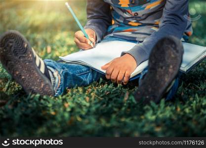Little child schoolkid writing on a notebook with pencil while sitting on green grass in the park. Kid education concept.