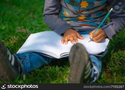 Little child schoolkid writing on a notebook with pencil while sitting on green grass in the park. Kid education concept.