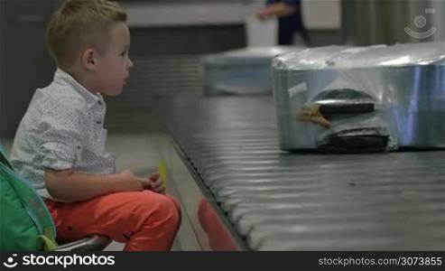 Little child sitting by the conveyor belt with luggage at the airport and watching people getting their bags