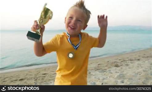 Little child with cup and medal jumping for joy to be the winner. He shouting and kissing prize at the seaside