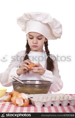 Little cook girl in a white apron breaks eggs in a deep dish, isolated on white background